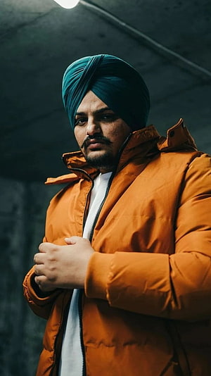 Portrait of a man wearing a teal turban and an orange puffer jacket standing in a dimly lit industrial setting, Sidhu Moose Wala, Indian Singer HD phone wallpaper