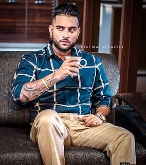 Karan Aujla in a teal checkered shirt and beige trousers, seated on a sofa holding a cup HD phone wallpaper
