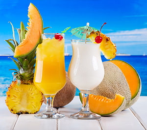 Tropical fruit cocktails with pineapple, cantaloupe, and coconut on a white table against a blue ocean backdrop HD wallpaper