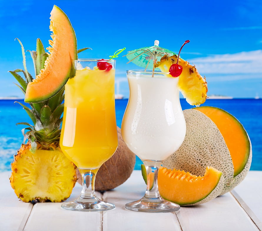 Tropical fruit cocktails with pineapple, cantaloupe, and coconut on a white table against a blue ocean backdrop HD wallpaper