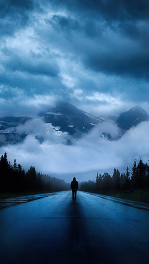 Lone Figure Walking on Wet Road Towards Misty Mountains Under Stormy Blue Sky HD phone wallpaper