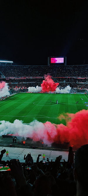 Vibrant red and white smoke flares illuminate a packed football stadium at night HD phone wallpaper