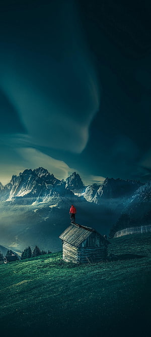 Lone Figure in Red Jacket Standing on Cabin Roof Overlooking Majestic Mountain Range HD phone wallpaper