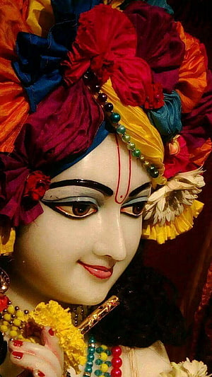 Close-up of Krishna deity adorned with vibrant floral headdress and traditional jewelry HD phone wallpaper
