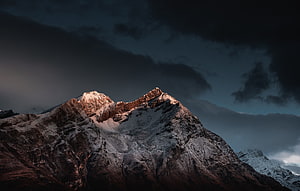 Dramatic Snow-Capped Mountain Peak Illuminated by Golden Hour Light Against Dark Stormy Sky HD wallpaper