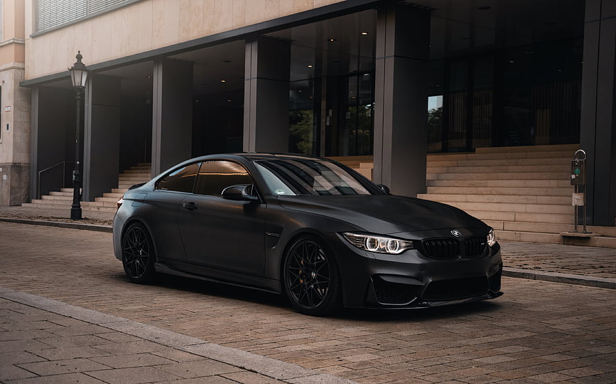 Matte Black BMW M4 Coupe Parked on Cobblestone Street in Front of Modern Building HD wallpaper