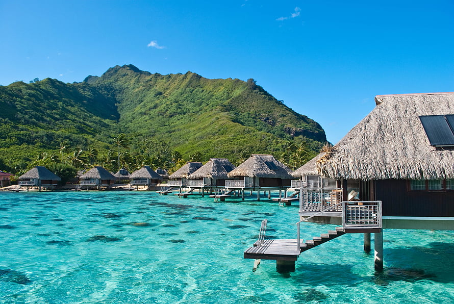 Overwater Bungalows With Thatched Roofs Set Against Lush Tropical Mountains And Turquoise Lagoon Waters HD wallpaper