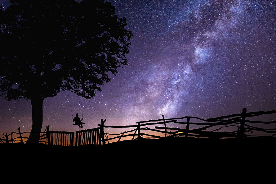 Silhouetted figure on a swing beneath the Milky Way and a rustic wooden fence HD wallpaper