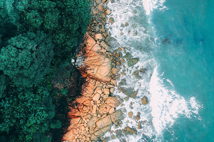 Aerial View of Turquoise Ocean Waves Crashing Against Rocky Shoreline and Dense Forest HD wallpaper