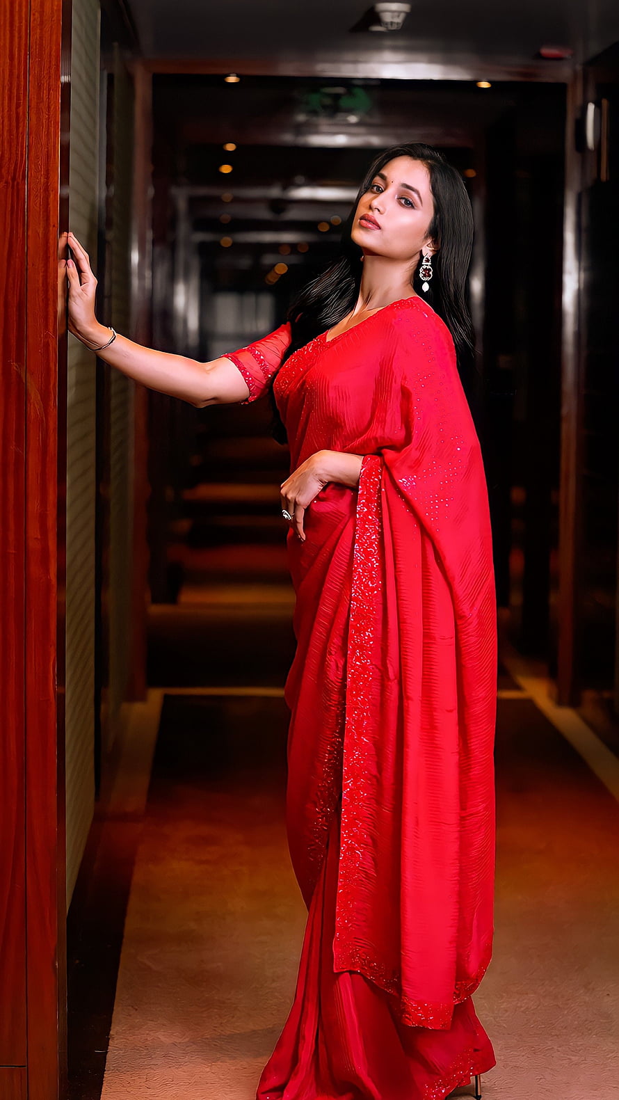 Srinidhi Shetty in a vibrant red embroidered saree posing in a dimly lit hotel corridor HD phone wallpaper