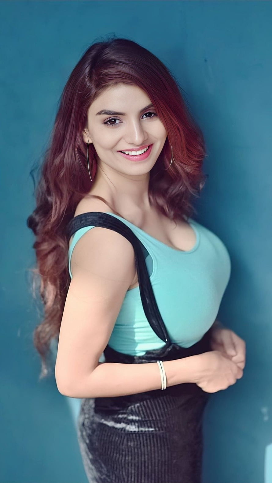 Portrait of a smiling woman with long wavy hair wearing a teal top and black skirt against a blue background, Anveshi jain, bollywood actress HD phone wallpaper