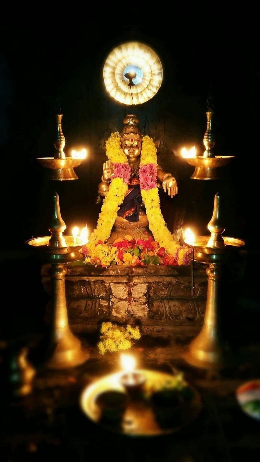 Golden Hindu deity statue adorned with marigold garlands and surrounded by lit brass oil lamps in a dark temple setting HD phone wallpaper