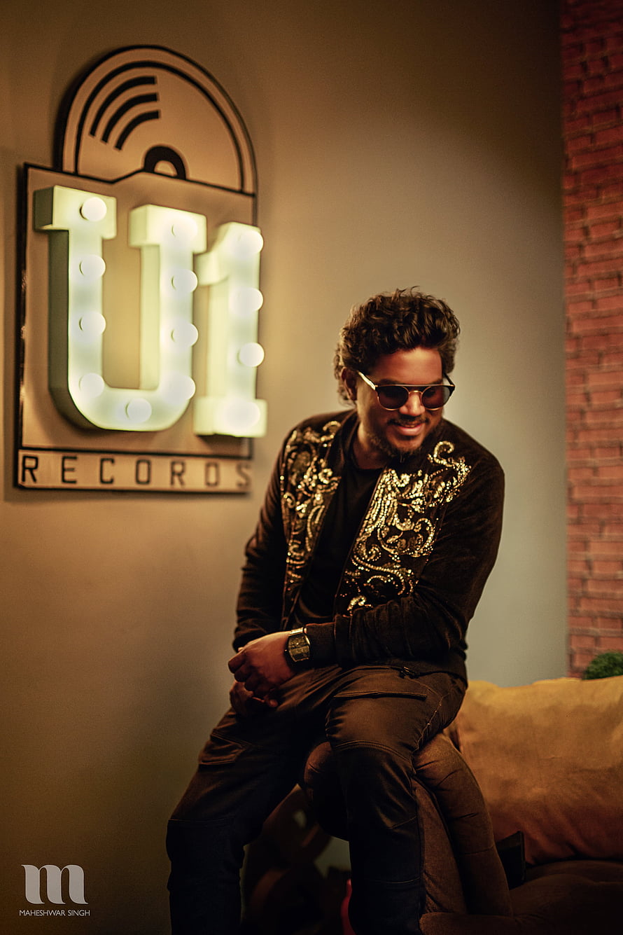 Yuvan Shankar Raja in a gold-embroidered jacket and sunglasses seated beside a U1 Records marquee sign HD phone wallpaper
