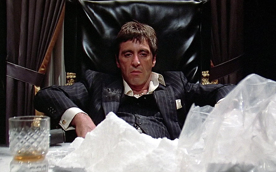 Tony Montana in a pinstripe suit reclining in a black leather chair with a glass of whiskey HD wallpaper