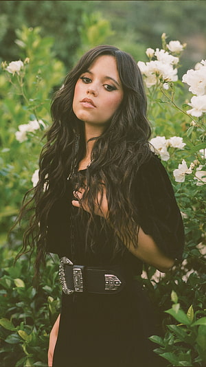 Jenna Ortega in a black dress with a silver belt standing among white roses in a garden HD phone wallpaper