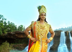 Krishna in golden attire standing confidently before a scenic waterfall backdrop HD wallpaper