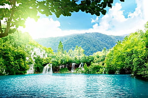Serene turquoise lake with cascading waterfalls framed by lush green forest and distant mountains HD wallpaper