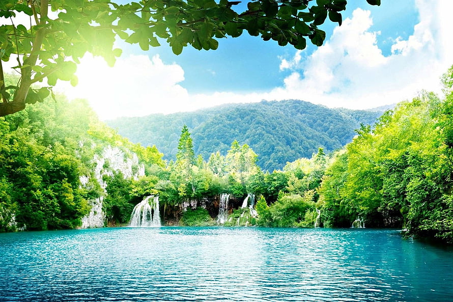 Serene turquoise lake with cascading waterfalls framed by lush green forest and distant mountains HD wallpaper