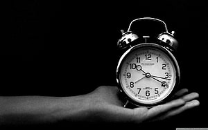Black and White Photograph of a Hand Holding a Classic Twin Bell Alarm Clock Against a Dark Background HD wallpaper