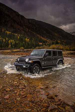 Jeep Wrangler fording a rocky mountain stream under a dramatic stormy sky HD phone wallpaper