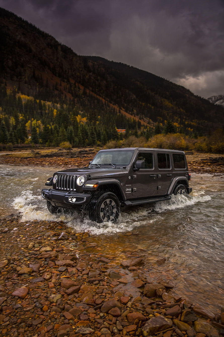 Jeep Wrangler fording a rocky mountain stream under a dramatic stormy sky HD phone wallpaper