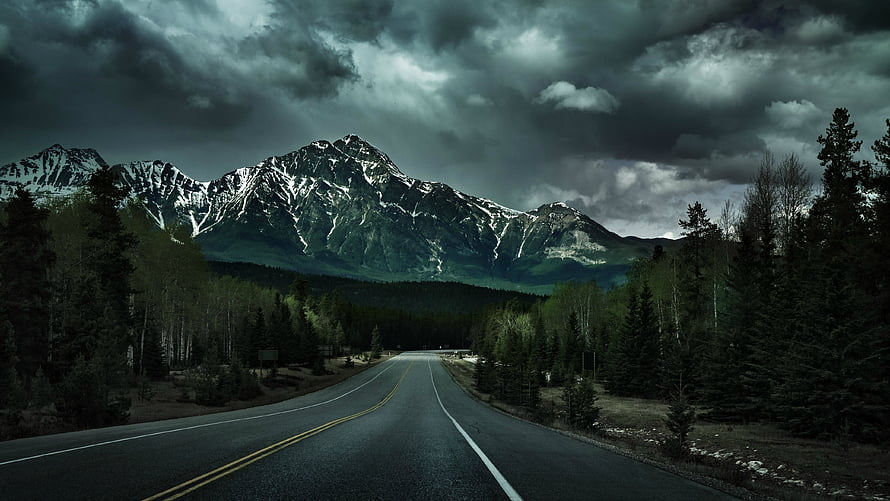 Moody Mountain Road Under Stormy Skies with Snow-Capped Peaks and Dense Forest HD wallpaper