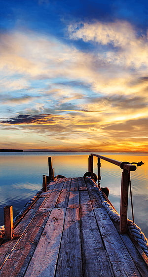 Weathered wooden pier extending into a calm lake under a vibrant golden sunset sky HD phone wallpaper