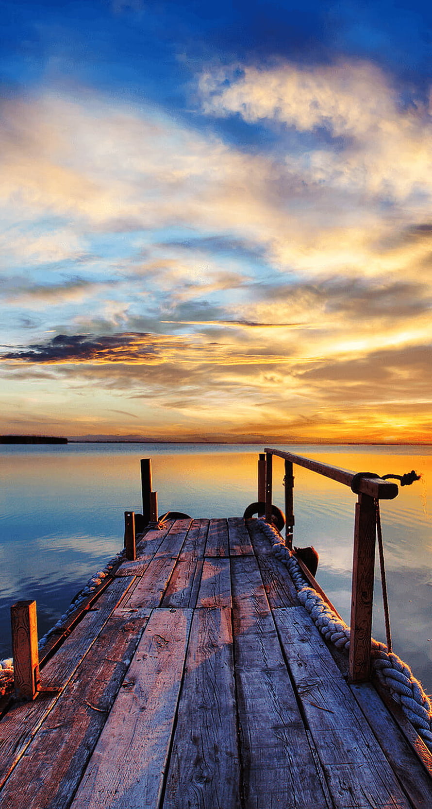 Weathered wooden pier extending into a calm lake under a vibrant golden sunset sky HD phone wallpaper
