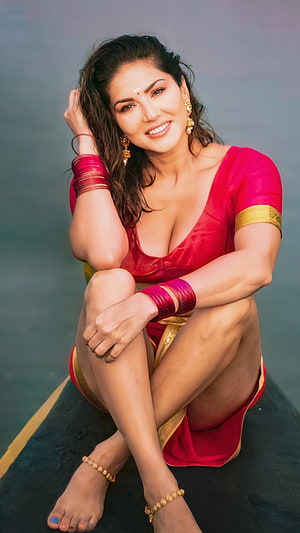 Sunny Leone in a vibrant red saree with gold jewelry posing by the water HD phone wallpaper