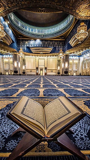 Open Quran on wooden stand inside grand mosque with blue carpet and golden dome HD phone wallpaper
