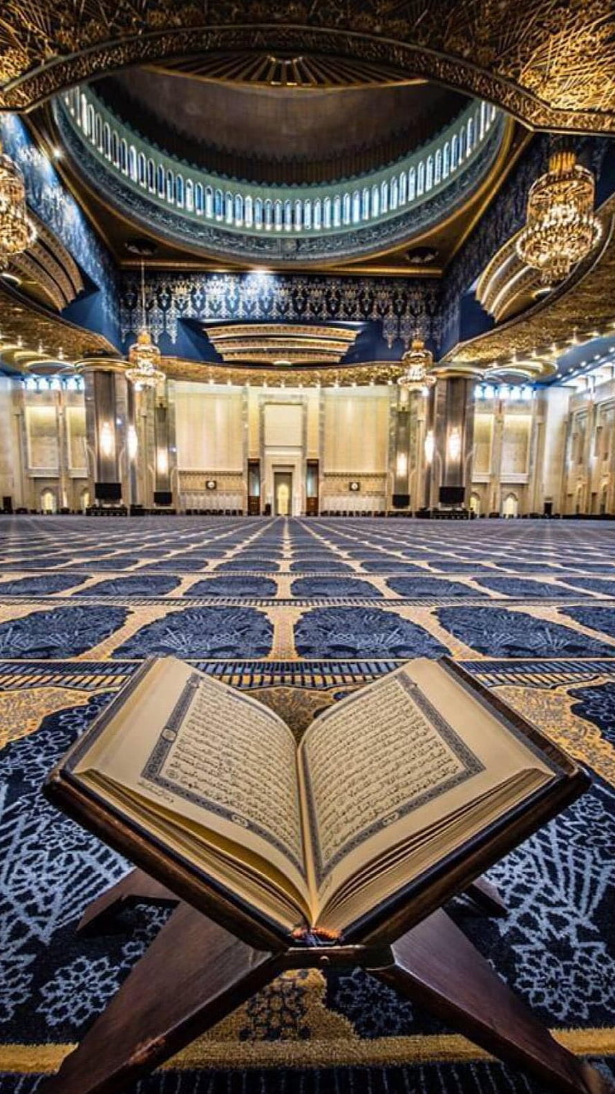 Open Quran on wooden stand inside grand mosque with blue carpet and golden dome HD phone wallpaper