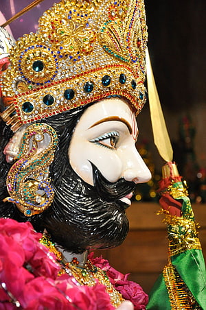 Close-up of a Hindu deity statue adorned with a jeweled golden crown, black beard, and pink flower garland HD phone wallpaper
