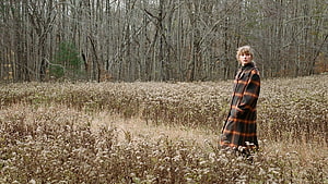 Taylor Swift in a plaid coat standing in a field of dried wildflowers with a bare forest in the background HD wallpaper