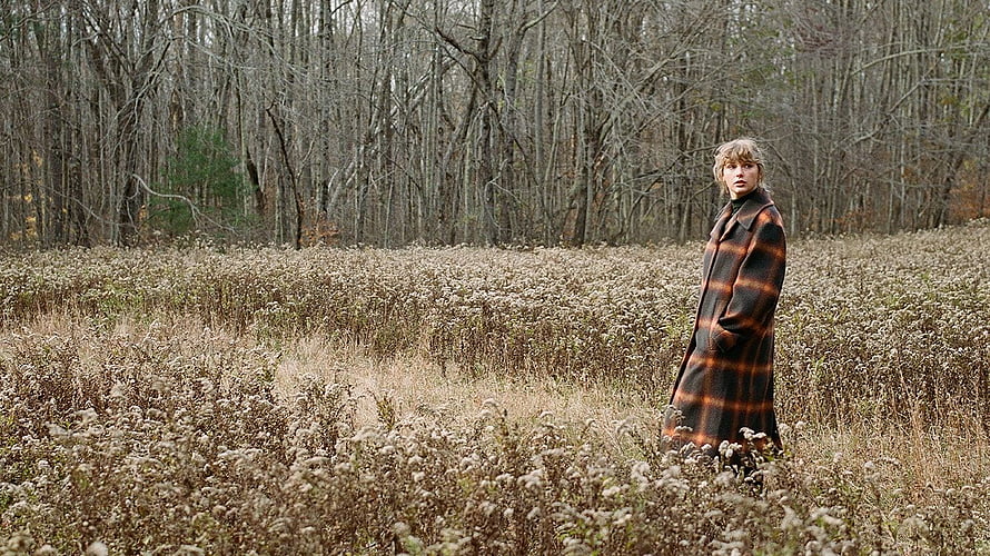 Taylor Swift in a plaid coat standing in a field of dried wildflowers with a bare forest in the background HD wallpaper