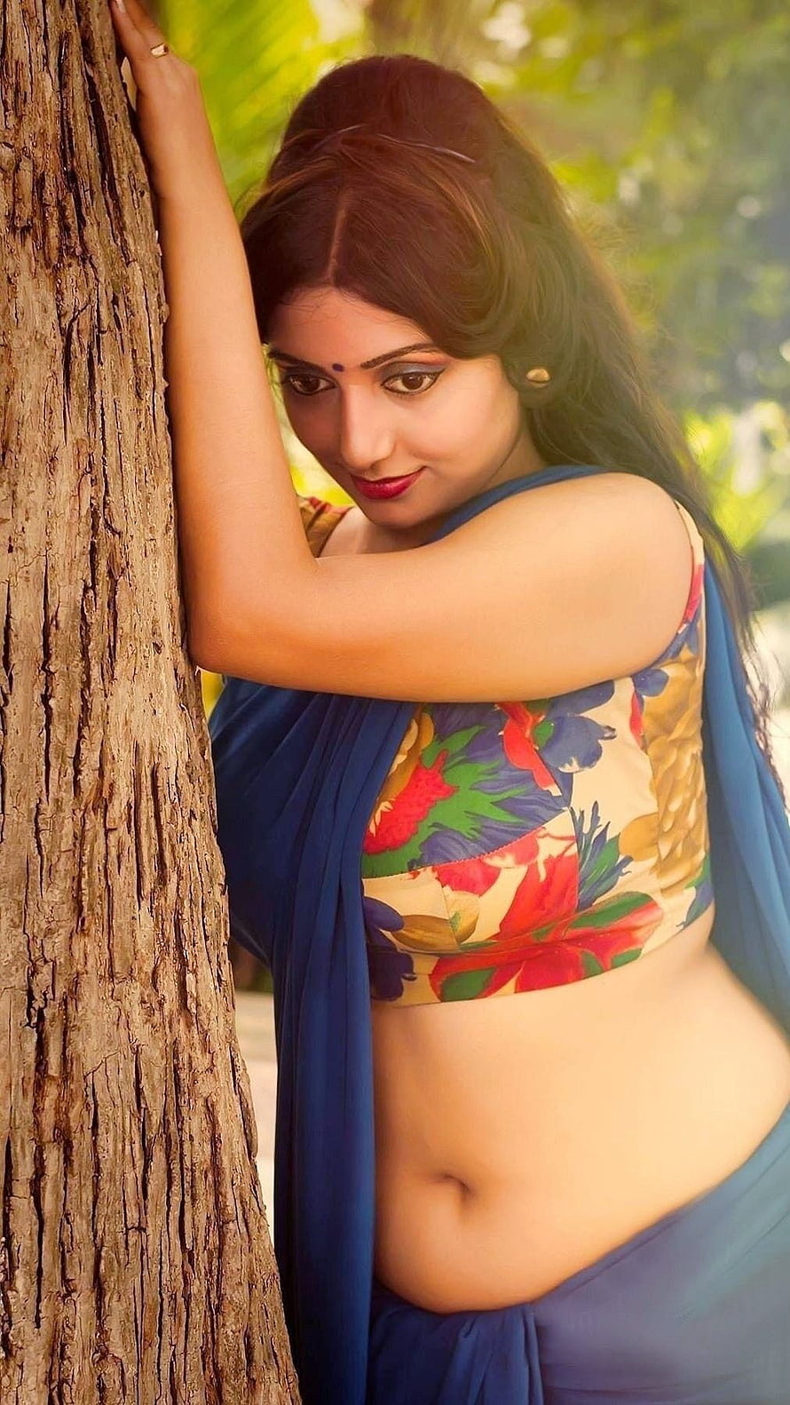 Rupsa Saha Choudhary in a floral blouse and blue saree leaning against a tree trunk in a garden HD phone wallpaper