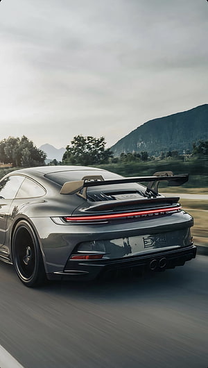 Grey Porsche 911 GT3 RS with large rear wing driving on a scenic mountain road HD phone wallpaper