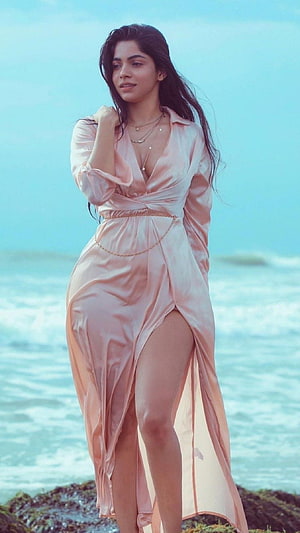 Divya Bharti in a flowing pink satin dress standing on a rocky shoreline with ocean waves behind her HD phone wallpaper