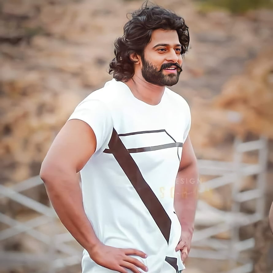Prabhas in white t-shirt with brown geometric design, smiling outdoors with rocky background HD phone wallpaper