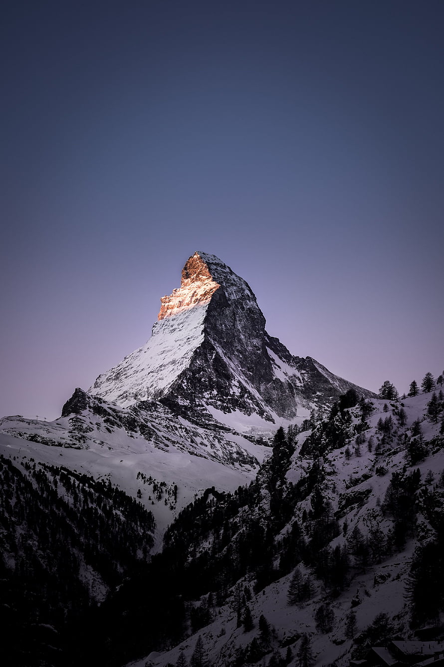 Matterhorn mountain peak illuminated by alpenglow against a twilight sky with snow-covered slopes and pine trees HD phone wallpaper