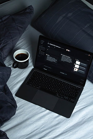 Black laptop on bed with coffee mug and dark blue pillows, cozy workspace setup HD phone wallpaper
