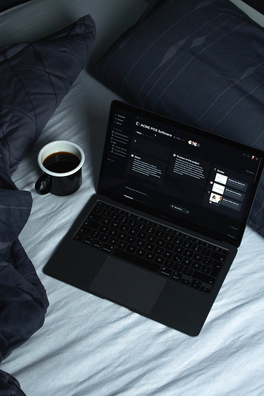 Black laptop on bed with coffee mug and dark blue pillows, cozy workspace setup HD phone wallpaper