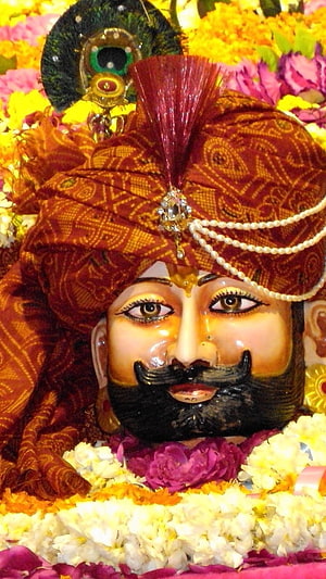 Close-up of a Hindu deity statue adorned with a red turban, peacock feather, and floral garlands HD phone wallpaper