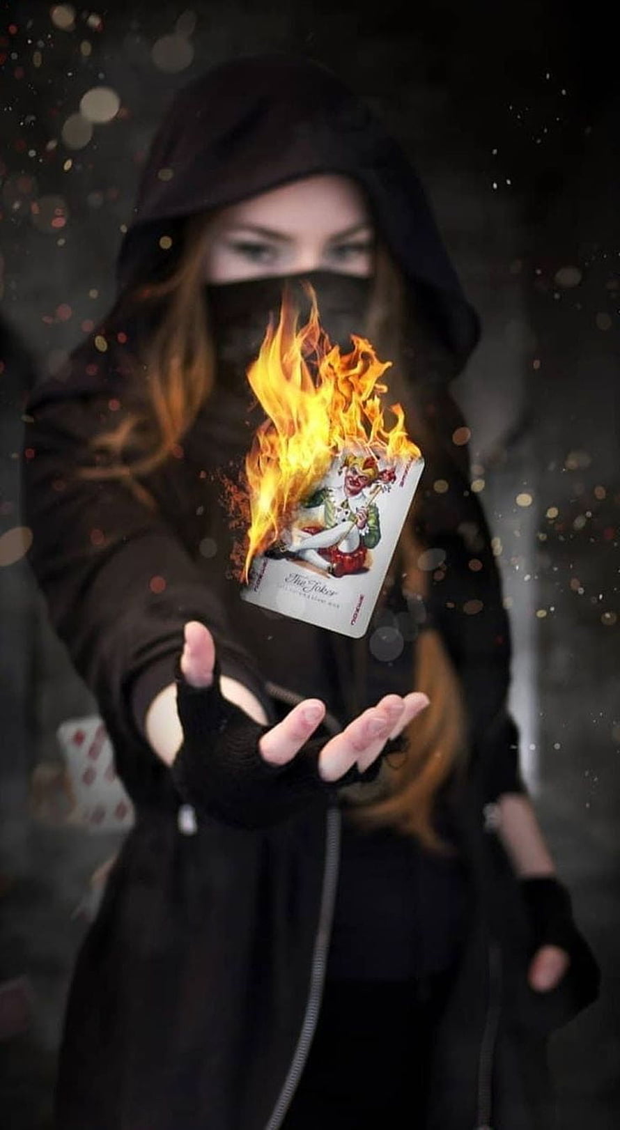 Mysterious hooded figure holding a burning joker playing card with magical fire effects HD phone wallpaper