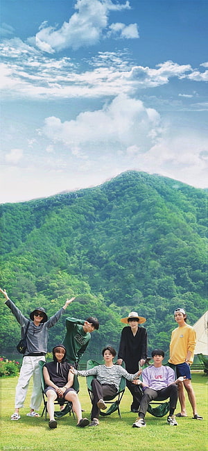 BTS Members Relaxing On A Grassy Lawn With Lush Green Mountains And Blue Sky In The Background HD phone wallpaper