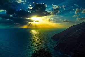 Dramatic Sunset Over Ocean With Sun Rays Breaking Through Clouds And Illuminating The Water Near A Rocky Cliff HD wallpaper