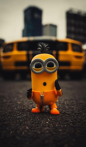 Cute Minion Toy Standing on Asphalt with Blurred Yellow Car Background HD phone wallpaper