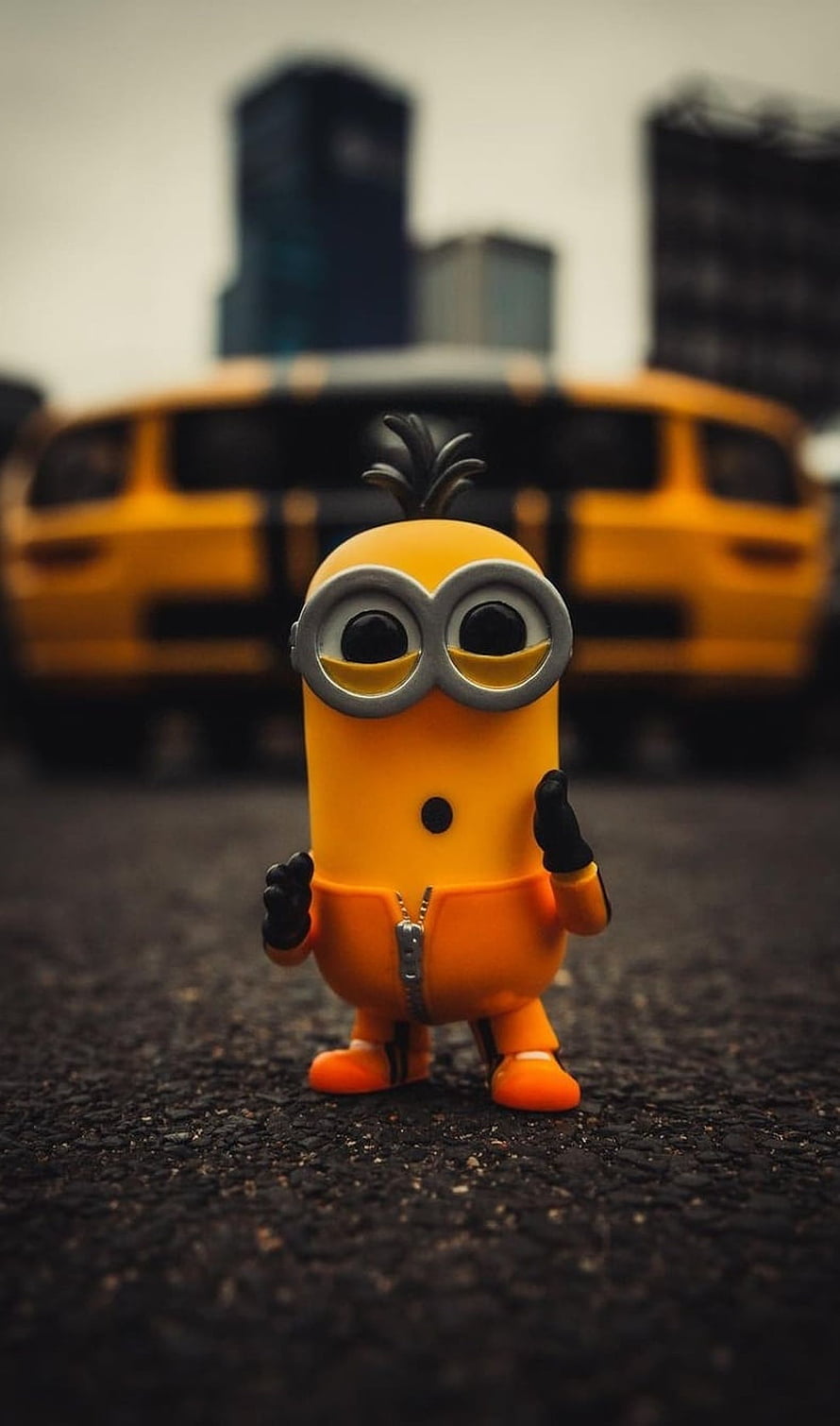 Cute Minion Toy Standing on Asphalt with Blurred Yellow Car Background HD phone wallpaper