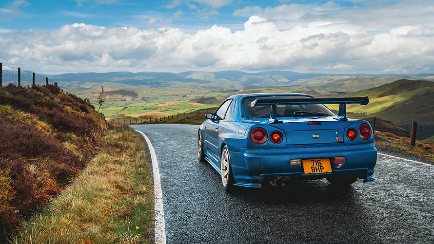 Blue Nissan Skyline GT-R R34 on a winding mountain road with scenic countryside backdrop HD wallpaper