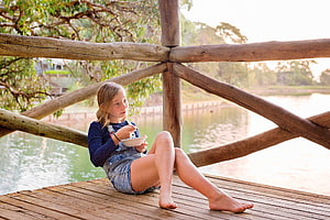 Barefoot girl eating on a wooden dock by a calm lake in a rustic outdoor setting HD wallpaper