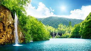 Serene turquoise lake surrounded by lush green forest and cascading waterfalls under a bright blue sky HD wallpaper